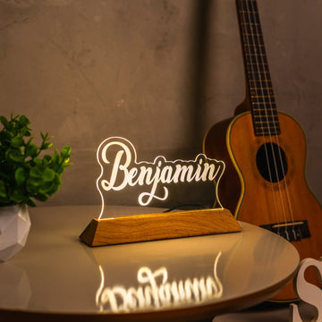 Luminária de acrílico personalizada com nome Benjamin sobre mesa, violão e planta ao fundo.