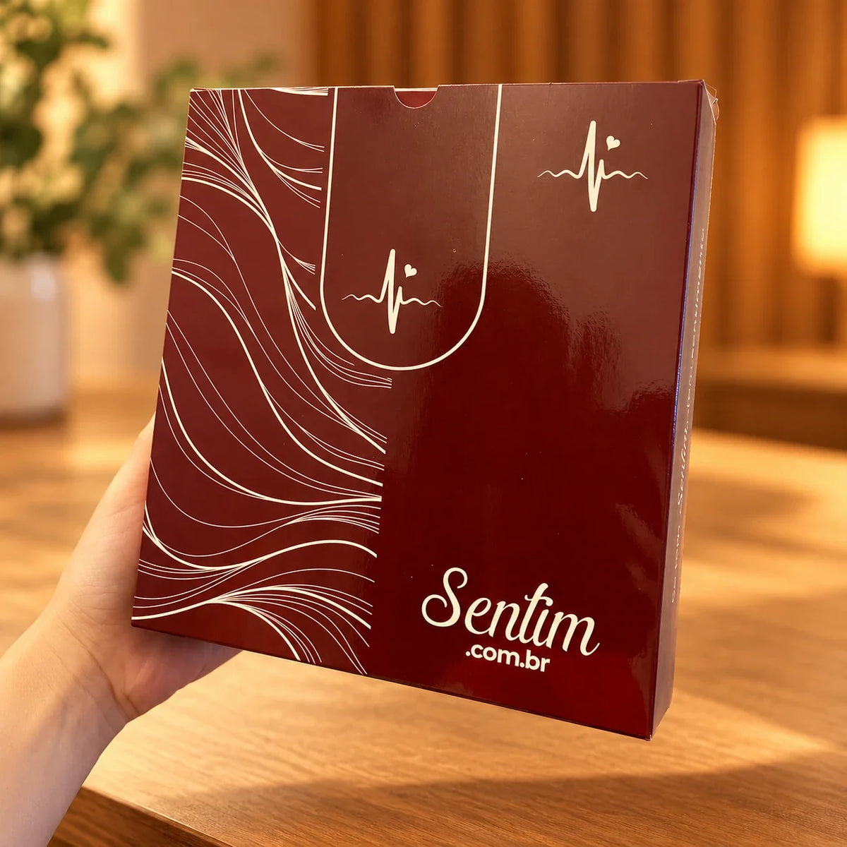 Caixa bordô da Sentim com o logo Sentim.com.br, segurada por uma mão sobre uma mesa de madeira