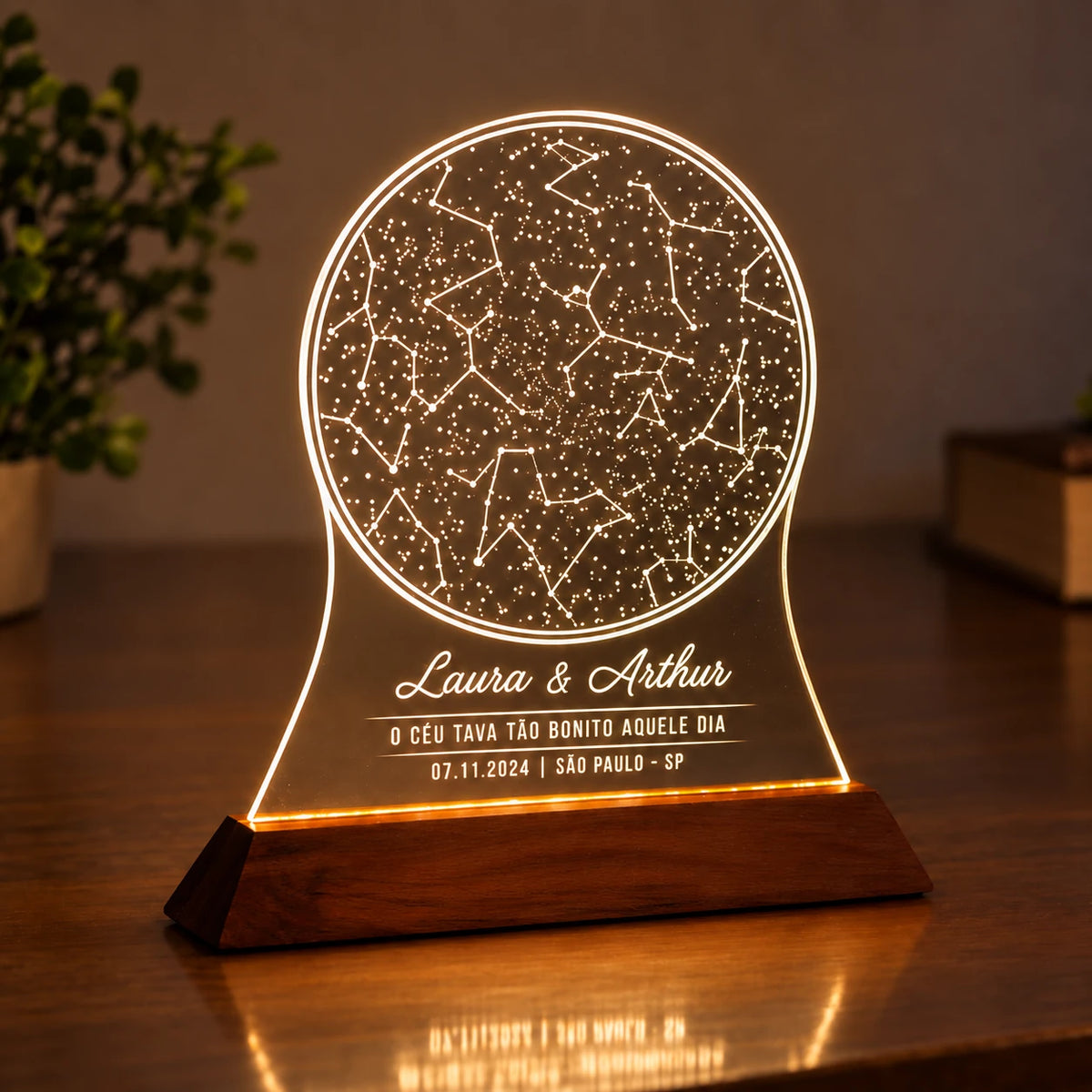 Luminária Mapa do Céu acesa, com mapa estelar e nomes Laura e Arthur no acrílico sobre base de madeira escura.