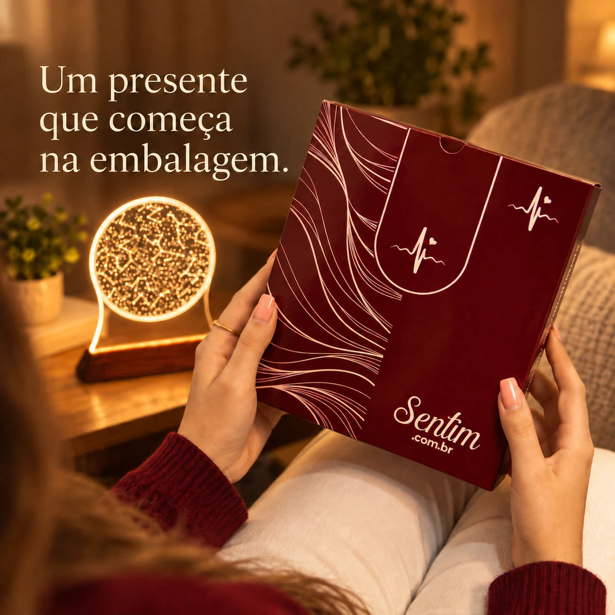 Caixa vinho da Sentim segurada nas mãos, com a luminária Mapa do Céu acesa ao fundo.