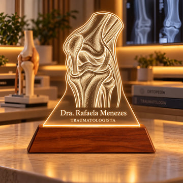 Luminária Traumatologista personalizada acesa, com desenho de joelho anatômico e o nome Dra. Rafaela Menezes gravados em acrílico sobre base de madeira